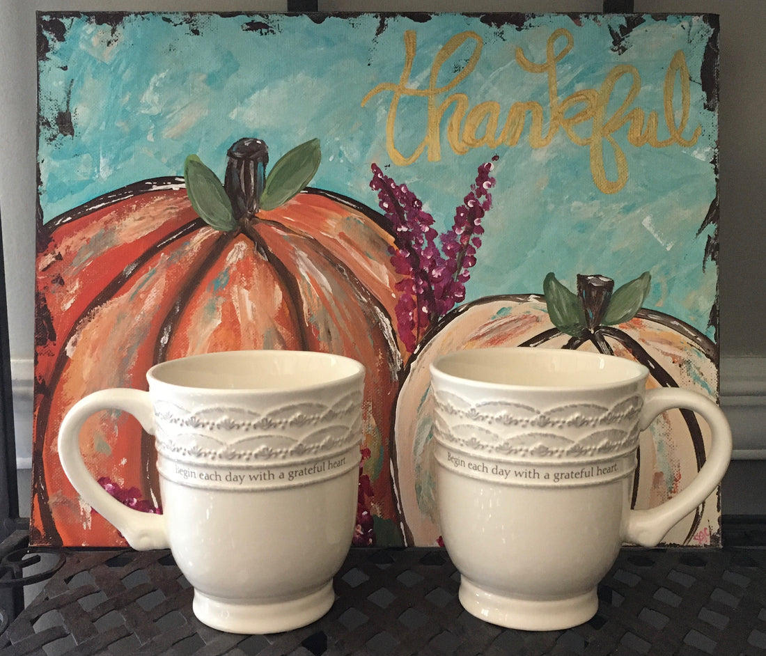 A painting of two pumpkins with the word "Thankful" on it and two mugs sitting in front of the painting.